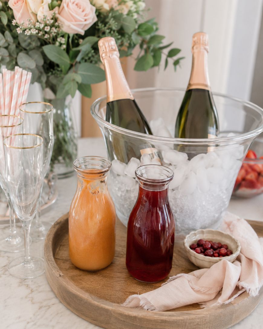 Three champagne bottles chill in ice beside glasses, while a tray holds two juice carafes—orange and red—a bowl of cranberries, and a napkin; flowers and striped straws are also visible.