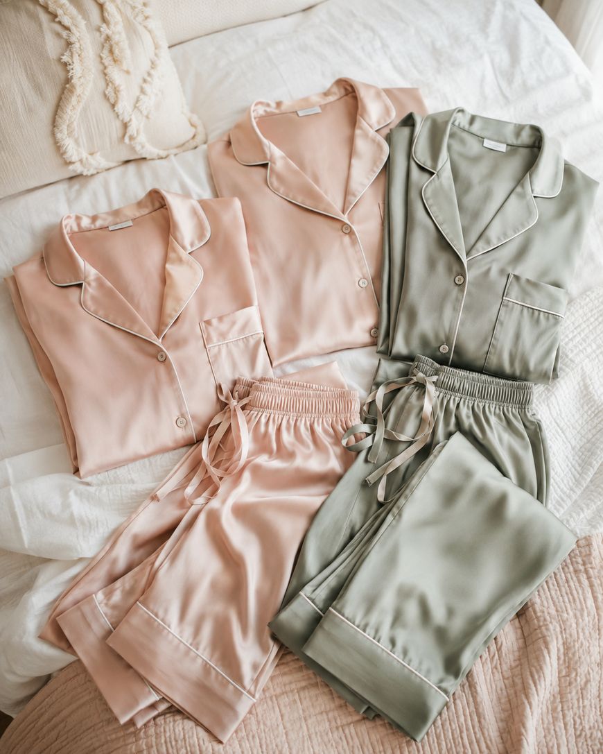 Two sets of satin pajamas, one light pink and one sage green, are neatly folded on a bed with cream bedding. Each set includes a button-up top and drawstring pants with contrasting piping detail.
