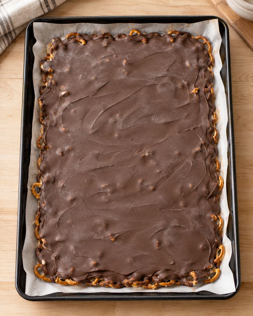 A large rectangular chocolate dessert topped with a smooth chocolate layer, with pretzel pieces visible around the edges, sits on parchment paper in a baking tray on a wooden surface.