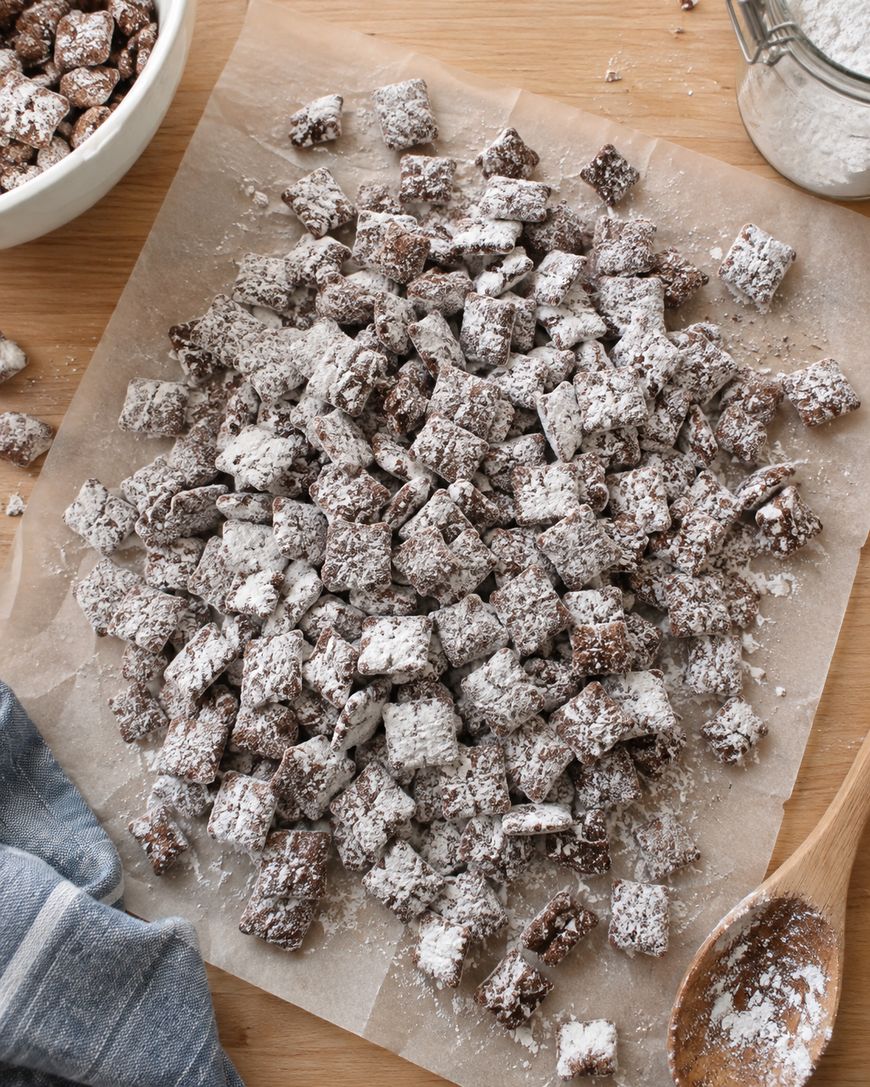 A pile of chocolate-covered cereal squares dusted with powdered sugar on parchment paper, with a wooden spoon, cloth, and a bowl of more cereal in the background.