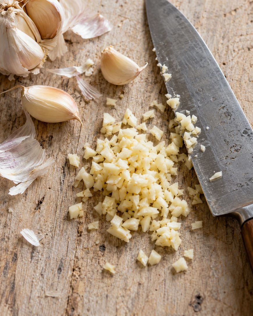 A pile of finely chopped garlic on a wooden surface, with a knife and garlic cloves nearby.