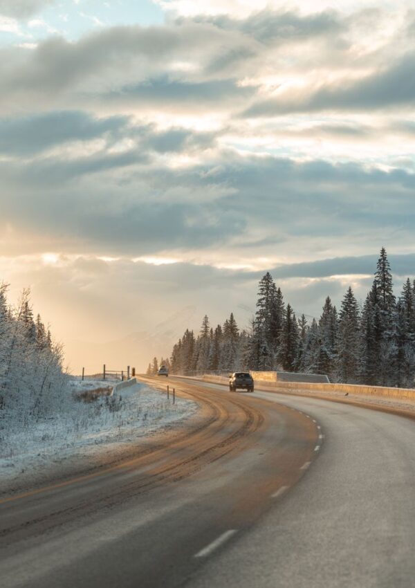 A car drives on a winding, snow-lined road through a winter landscape with tall pine trees and a dramatic sky at sunrise or sunset.