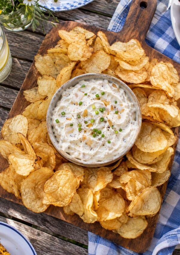 A bowl of creamy dip garnished with chopped chives is surrounded by potato chips on a wooden board, set on an outdoor table with a blue checkered cloth.