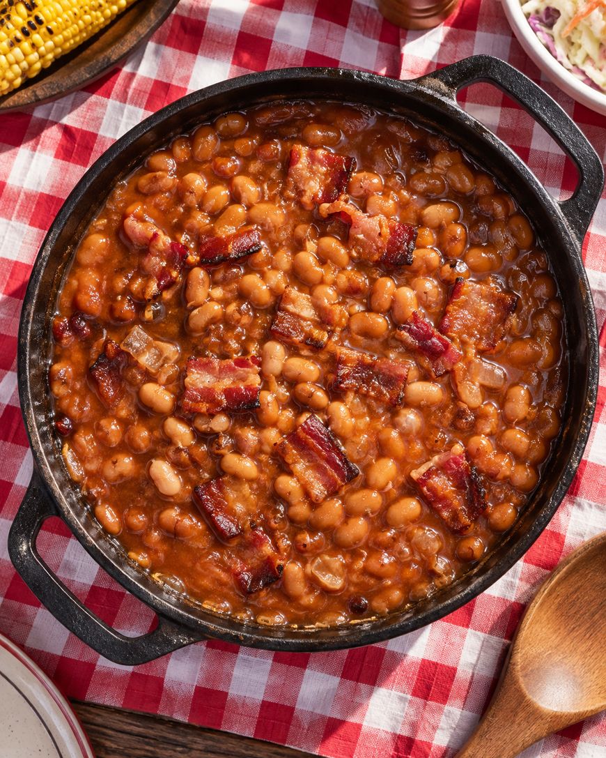A pot of baked beans topped with pieces of bacon sits on a red-and-white checkered tablecloth, next to a bowl of coleslaw and an ear of corn.