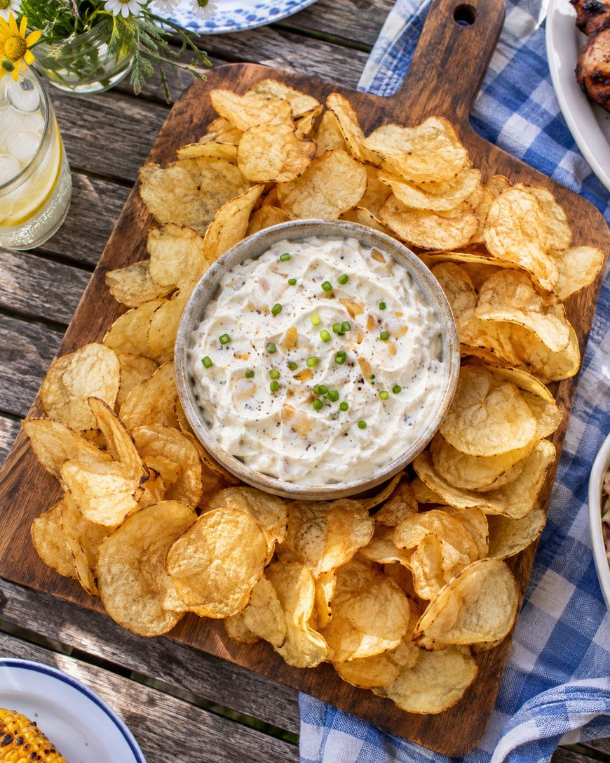A bowl of creamy dip garnished with chopped chives is surrounded by potato chips on a wooden board, set on a picnic table with a blue checkered cloth.