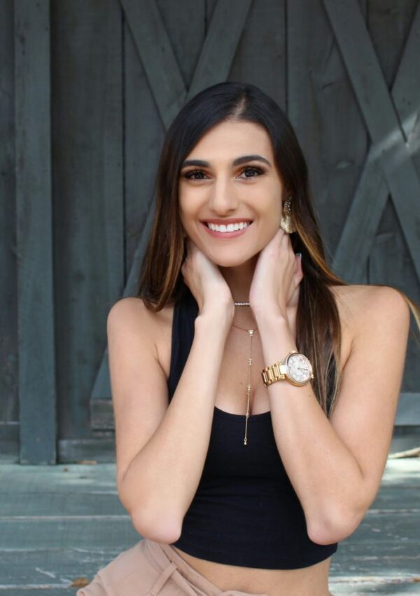 A woman with long brown hair smiles, holding her hands to her neck. She wears a black sleeveless top, gold watch, earrings, and layered necklaces. The background features a wooden wall with a rustic, gray finish.