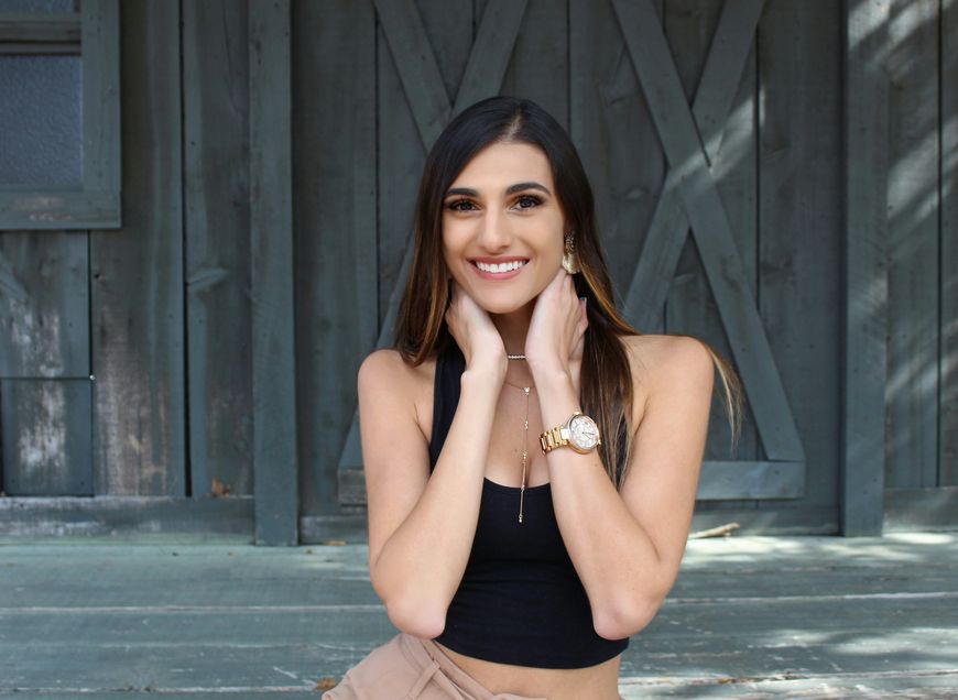A young woman with long dark hair, wearing a black sleeveless top and gold watch, smiles while holding her hands near her neck, standing in front of a rustic wooden wall.
