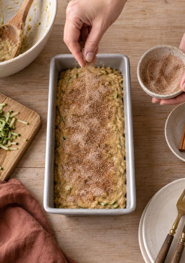 A person sprinkles cinnamon sugar over zucchini bread batter in a loaf pan, surrounded by bowls, cinnamon sticks, shredded zucchini, and utensils on a wooden table.