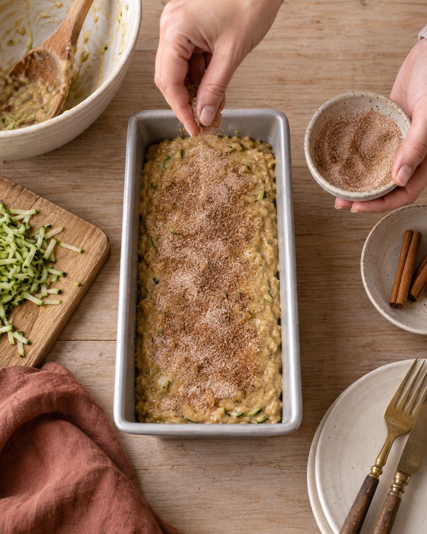 A person sprinkles cinnamon sugar on unbaked zucchini bread batter in a loaf pan. Grated zucchini, a bowl, plates, and utensils are arranged nearby on a wooden table.