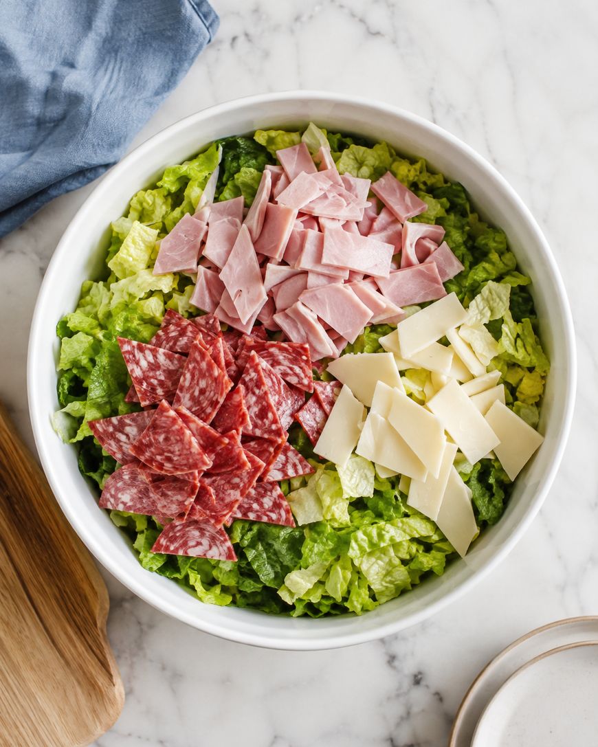 A white bowl filled with chopped romaine lettuce, topped with piles of sliced ham, salami, and strips of white cheese. The bowl is on a marble surface next to a blue napkin and a wooden board.