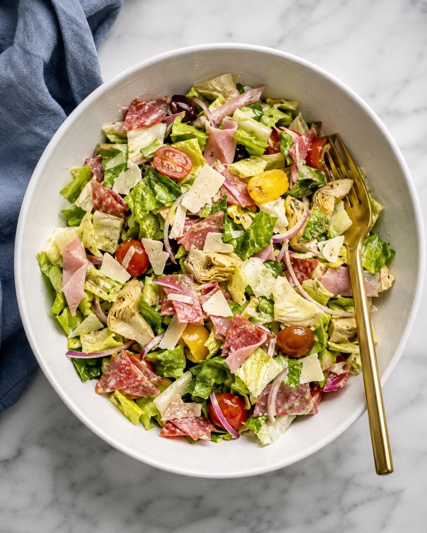 A bowl of chopped salad with lettuce, cherry tomatoes, salami, ham, artichoke hearts, red onions, and shaved parmesan, with a gold fork on the side and a blue napkin nearby.