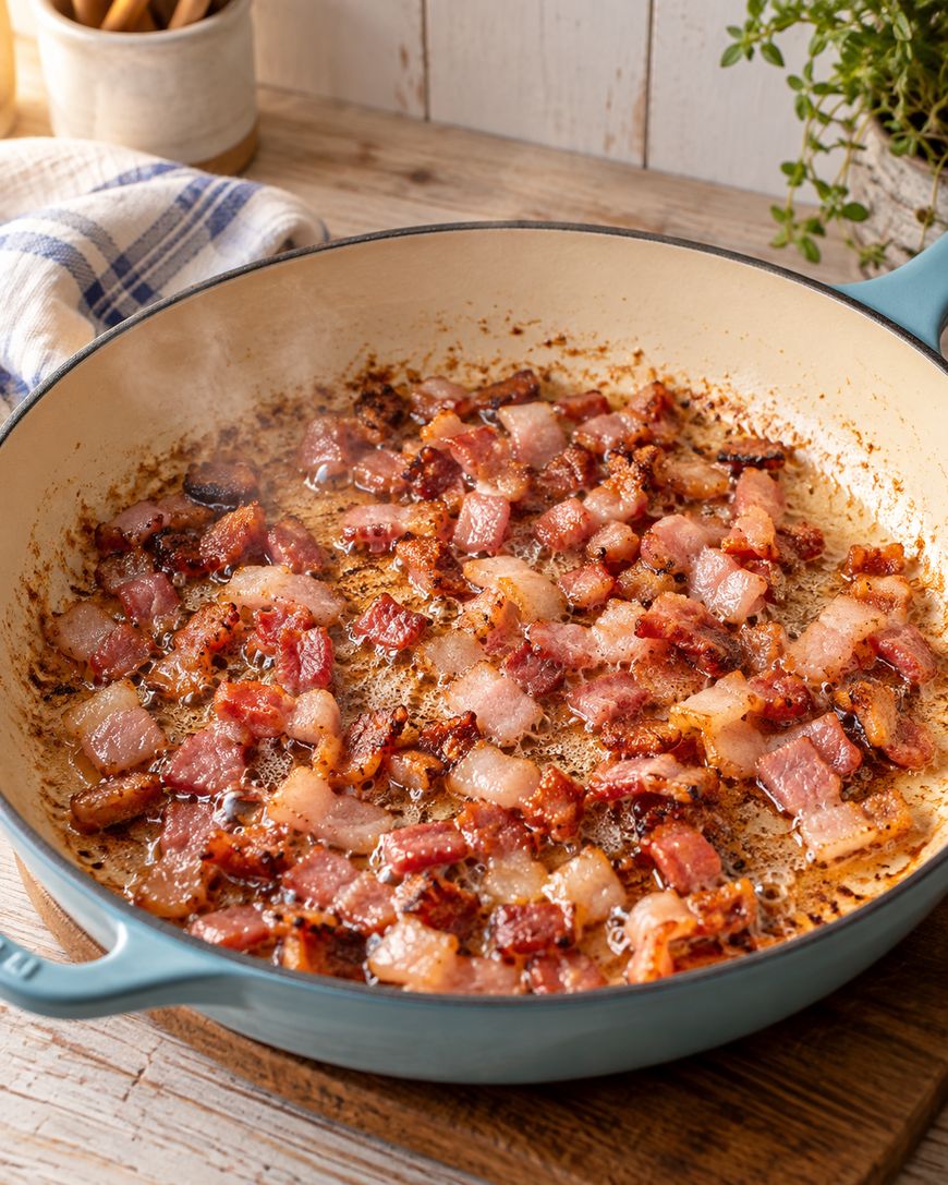 Diced bacon sizzling and browning in a large, light blue skillet on a wooden surface, with steam rising and a kitchen towel and herbs in the background.