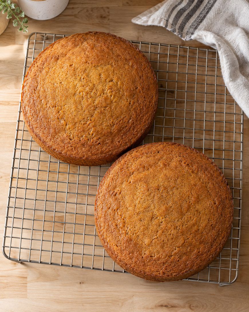 Two round, golden-brown cakes cooling on a wire rack atop a wooden surface, with a striped kitchen towel and a plant partially visible in the background.