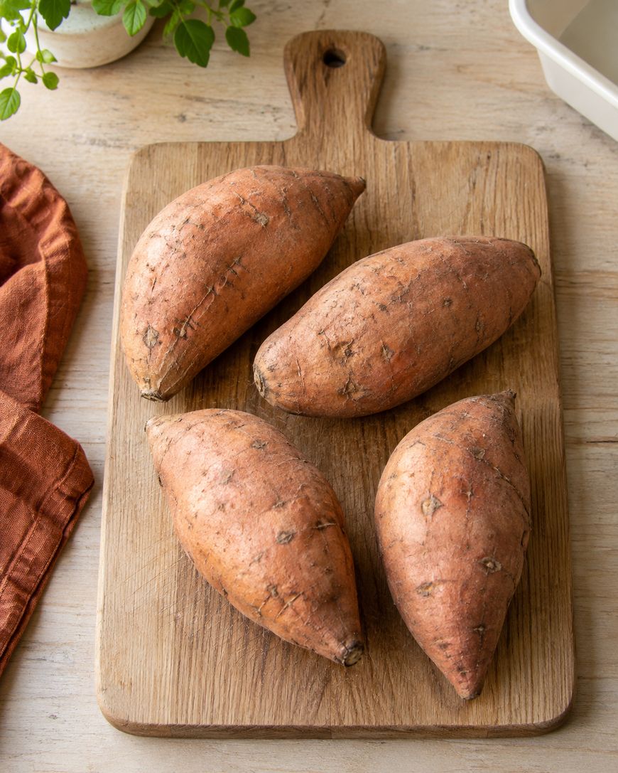 Four whole sweet potatoes with brownish skin rest on a wooden cutting board, which sits on a light wood surface. An orange cloth and green plant are partially visible nearby.