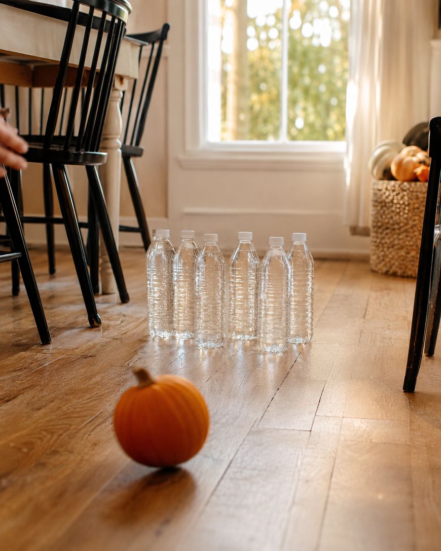 A small pumpkin on a wooden floor is aimed at a row of empty water bottles set up like bowling pins. Sunlight streams through a window in the background.