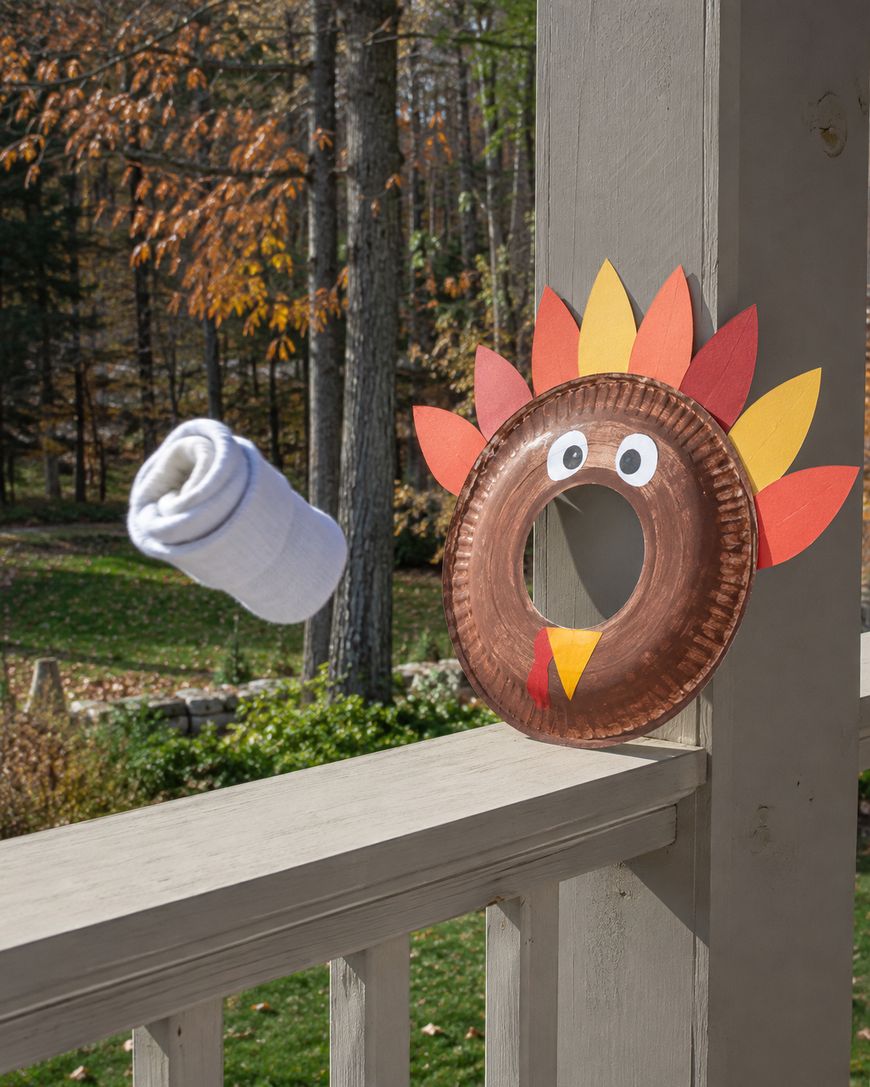 A paper plate decorated as a turkey is attached to a porch railing. A rolled-up sock is mid-air, aimed at the hole in the plate. Trees with autumn leaves are visible in the background.