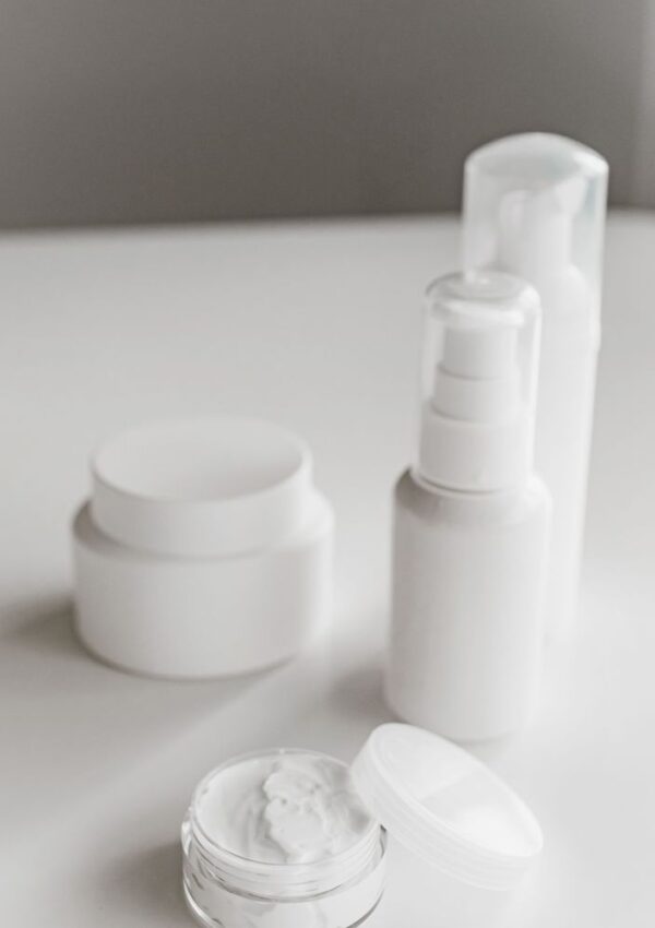 Four white cosmetic containers, including jars and pump bottles, are arranged on a white surface. One jar is open, revealing a cream inside. The scene has a minimalistic, clean aesthetic with soft lighting.