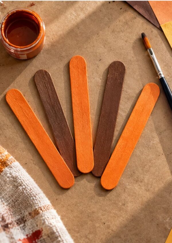 Five painted craft sticks in orange and brown shades are arranged in a fan shape on a table with a paintbrush, open paint container, and pieces of colored paper nearby.
