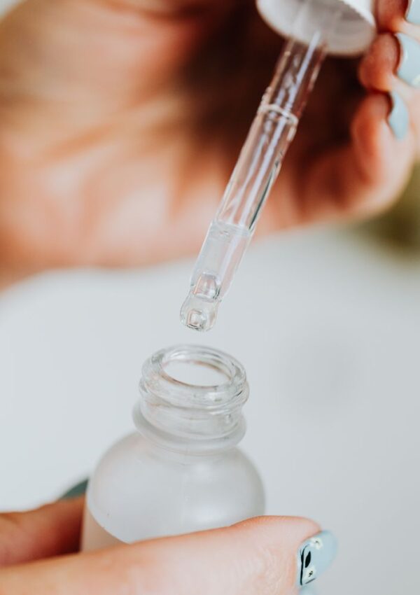 A hand holds a glass dropper above an open frosted bottle, about to dispense a clear liquid. Fingernails are painted light blue with a black pattern. The scene suggests skincare or cosmetic product use.