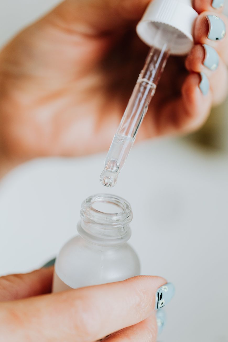 A hand holds a glass dropper above an open frosted bottle, with another hand steadying the bottle. The dropper is about to dispense liquid. The person has light blue painted nails.