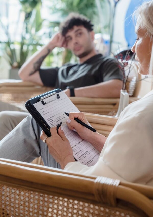 A person with a clipboard writes notes while another person sits across from them, appearing thoughtful. They are seated in wicker chairs in a bright room with plants.