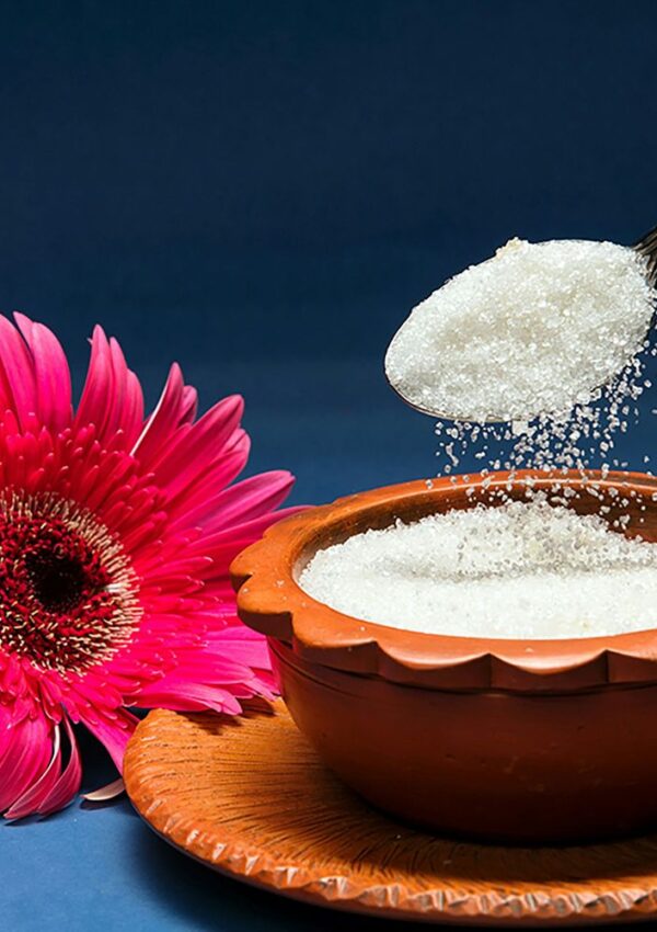 A spoon pouring white sugar into a clay bowl, next to a pink gerbera daisy.