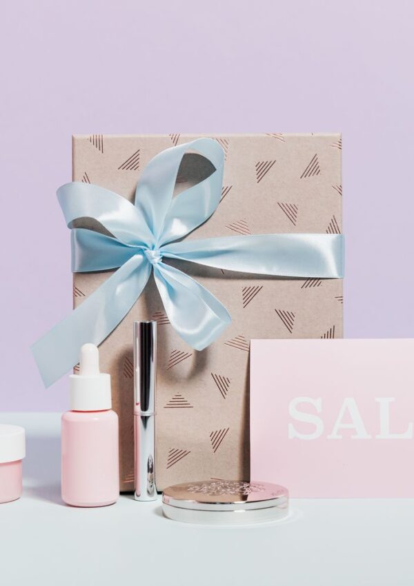 A gift box tied with a light blue ribbon, surrounded by beauty products and a card that says SALE.