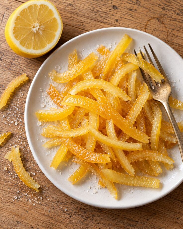 A pile of candied lemon peel on a small plate with a fork and a lemon half on a wooden table.