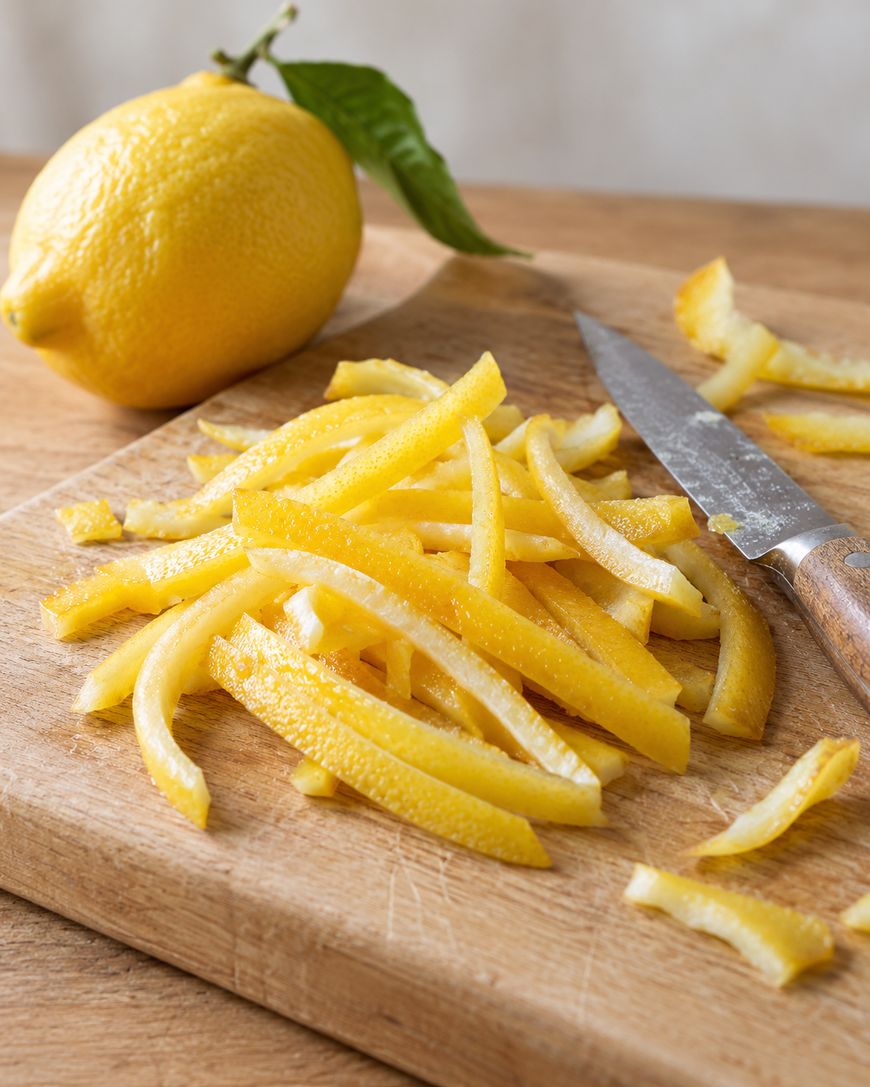 A whole lemon next to a pile of thin lemon zest strips and a paring knife on a wooden cutting board.