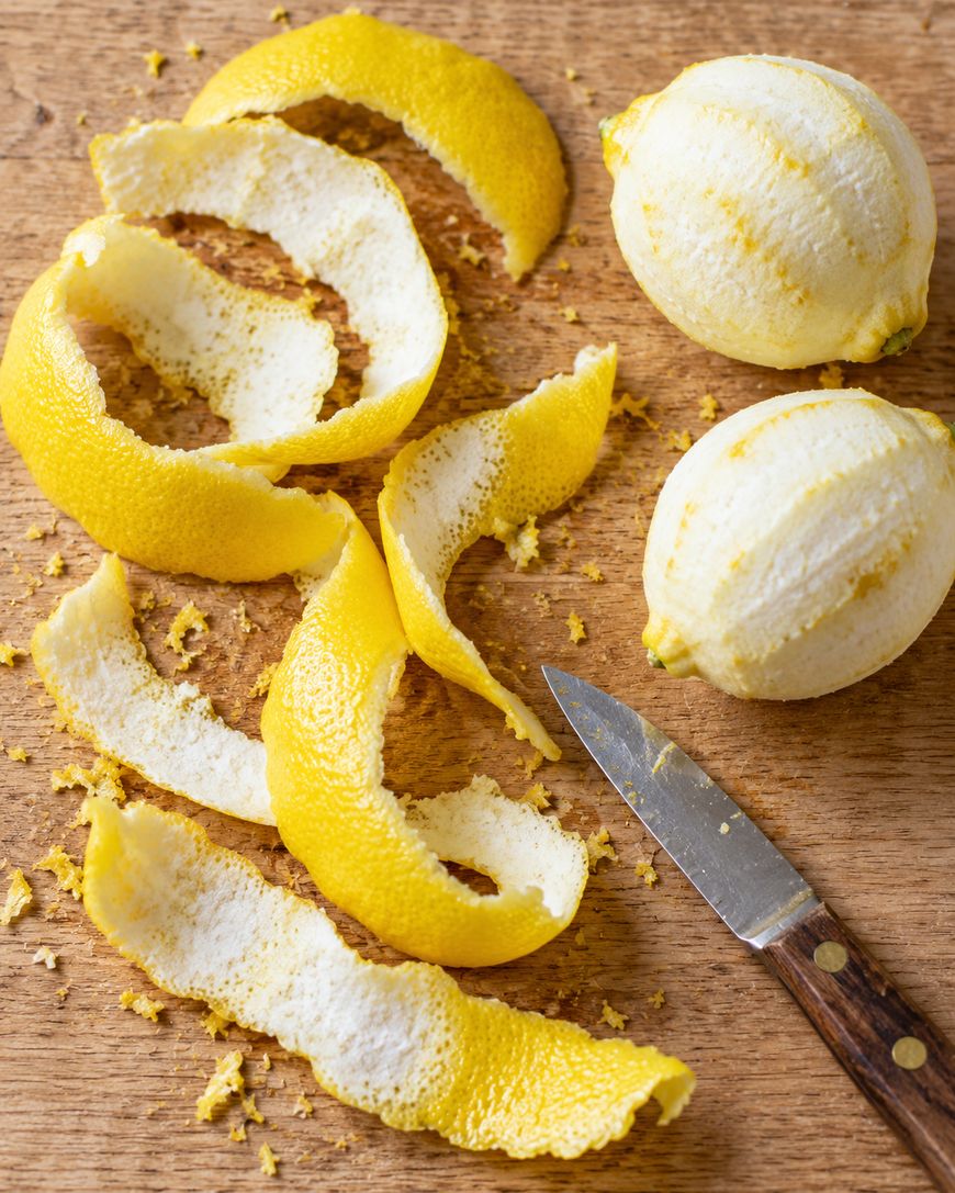 Two peeled lemons, lemon peels, and a paring knife on a wooden cutting board.