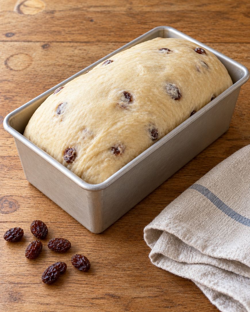 A loaf of unbaked raisin bread dough in a metal loaf pan, with a few loose raisins on a wooden surface.