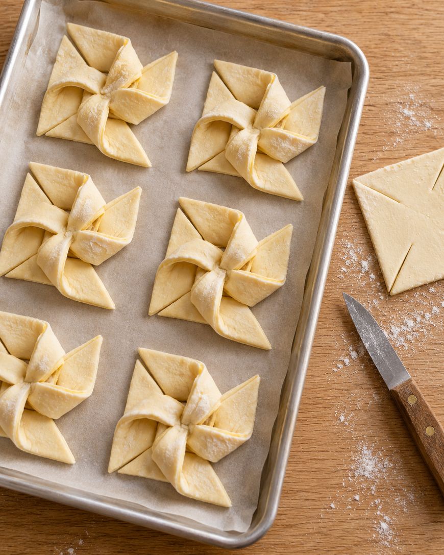 Five unbaked puff pastry pinwheels on a parchment-lined baking sheet, with one more pastry square and a paring knife on the side.