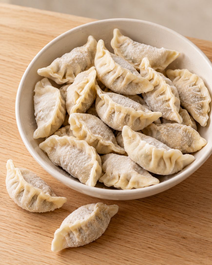 A bowl filled with raw, pleated dumplings on a wooden surface, with two additional dumplings resting outside the bowl.
