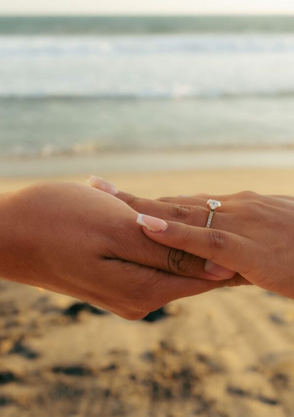 A person wearing a diamond engagement ring holds another persons hand at the beach.