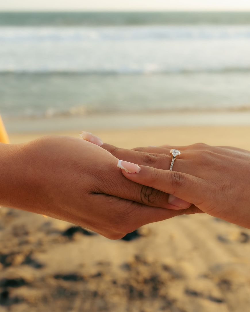 Two hands held together on a sunny beach with a diamond engagement ring on one finger