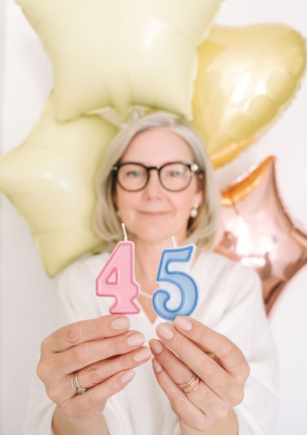 Woman with gray hair and glasses holding birthday candles shaped like the number 45 in front of pastel balloons