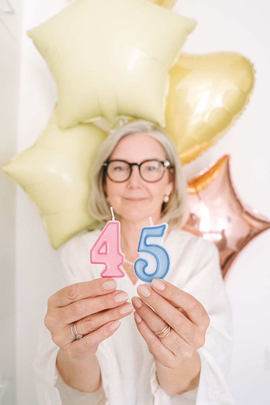 Woman with gray hair and glasses holding birthday candles shaped like the number 45 in front of pastel balloons