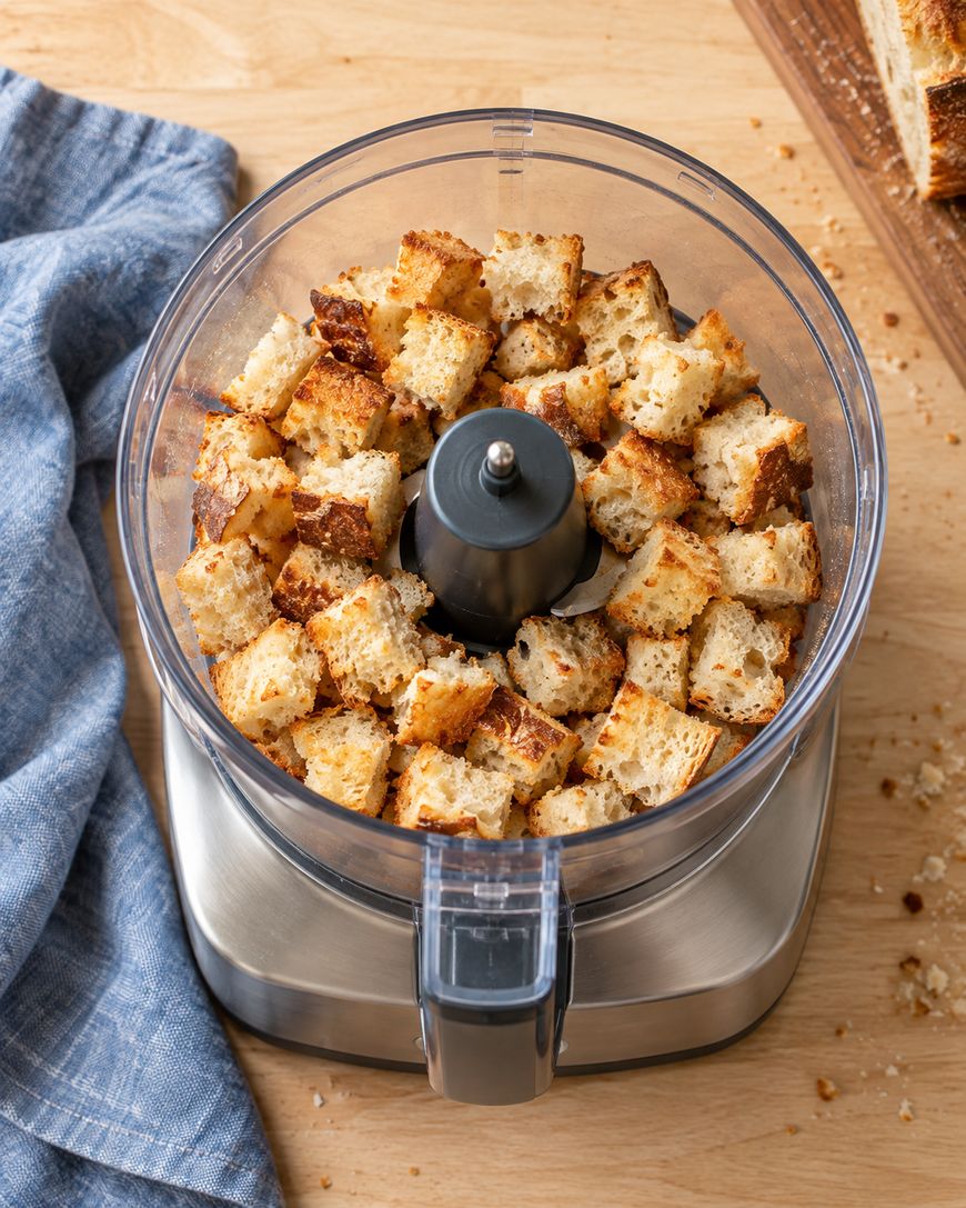 Cubed bread pieces inside a food processor on a wooden surface, with a blue cloth and more bread on a cutting board nearby.