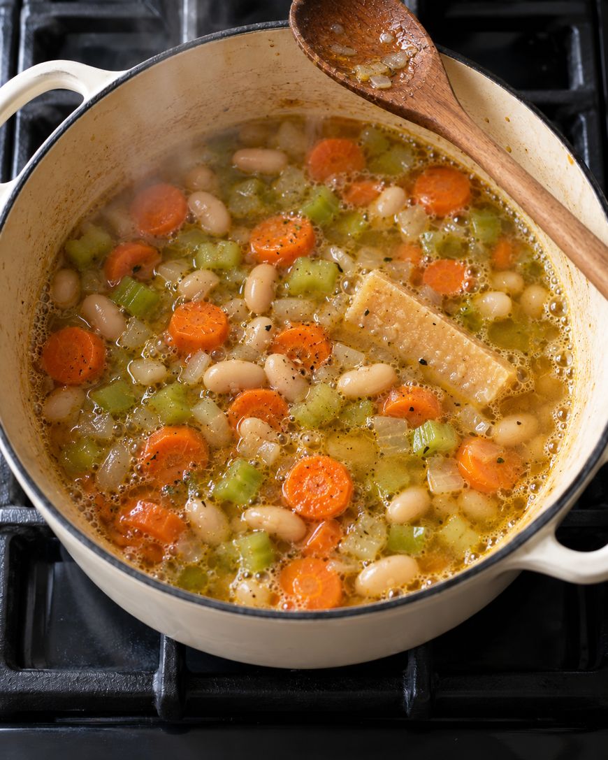 A white enameled cast iron pot on a stovetop filled with bean, carrot, and celery soup and a wooden spoon.