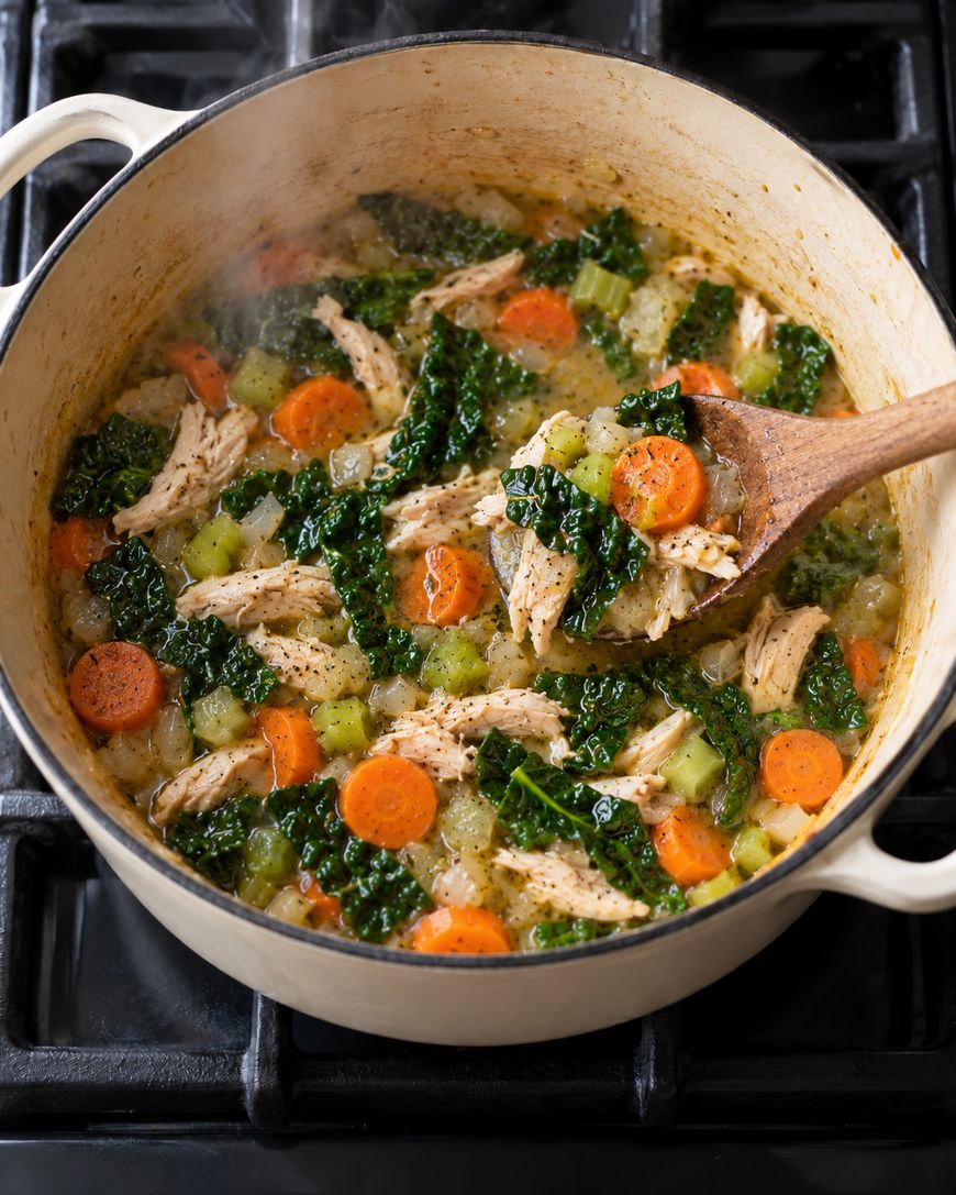 A wooden spoon lifts chicken, kale, and sliced carrots from a pot of simmering soup on a stovetop.