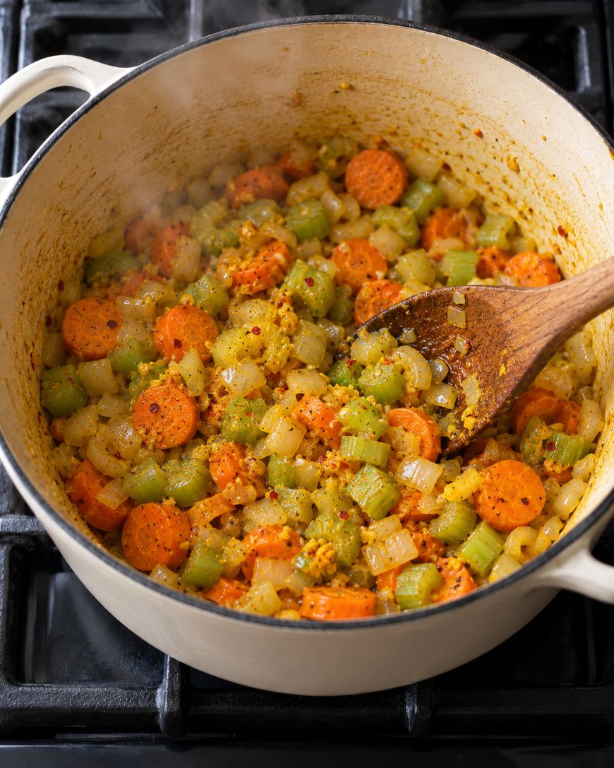 A wooden spoon stirs chopped carrots, celery, and onions sautéing in a large white enameled pot on a stovetop.