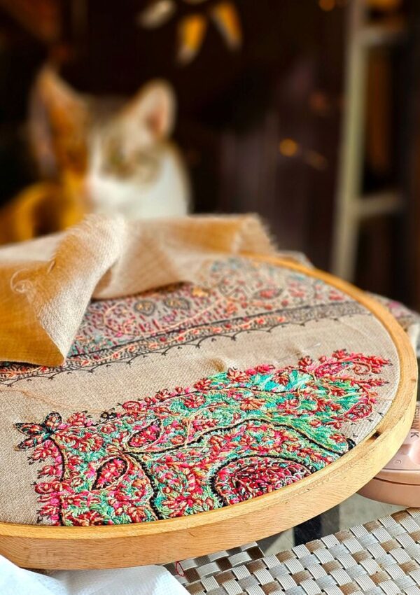 Embroidery hoop holding a fabric panel featuring intricate, colorful floral needlework, with a cat out of focus in the background.