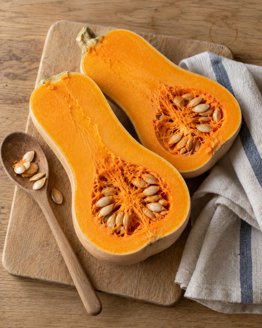A butternut squash cut in half lengthwise on a wooden cutting board with a wooden spoon and scattered seeds.