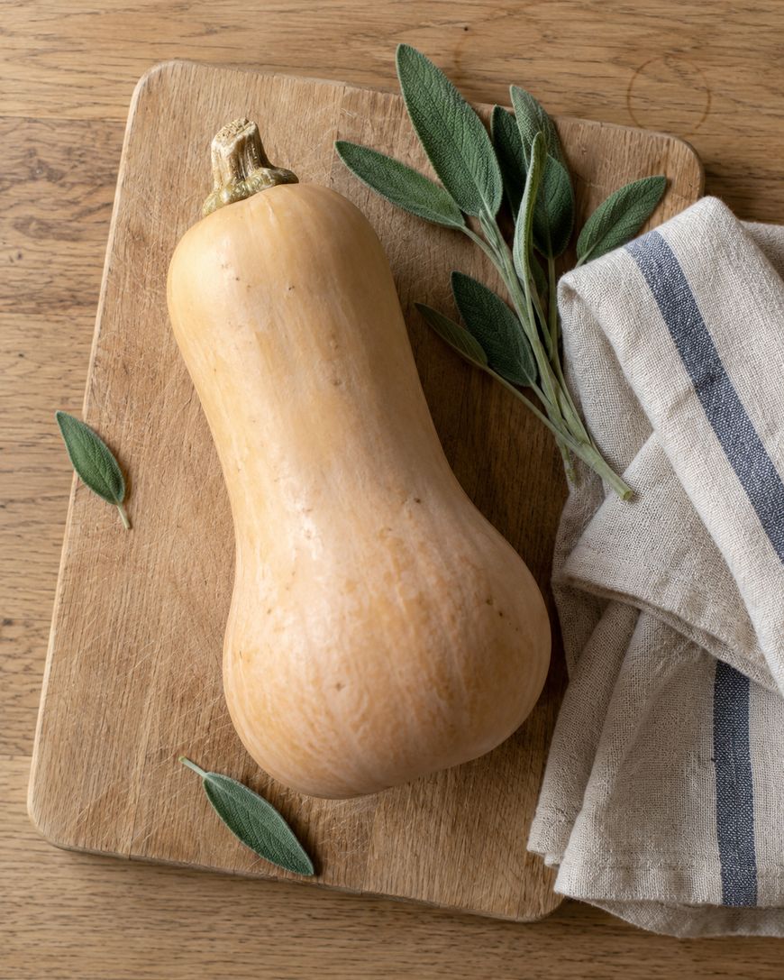 A butternut squash with fresh sage leaves on a wooden cutting board next to a striped kitchen towel.