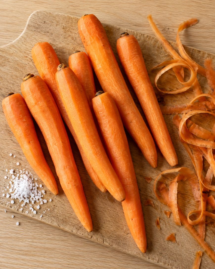 Several peeled carrots and a pile of carrot peelings on a wooden cutting board with a small heap of coarse salt.