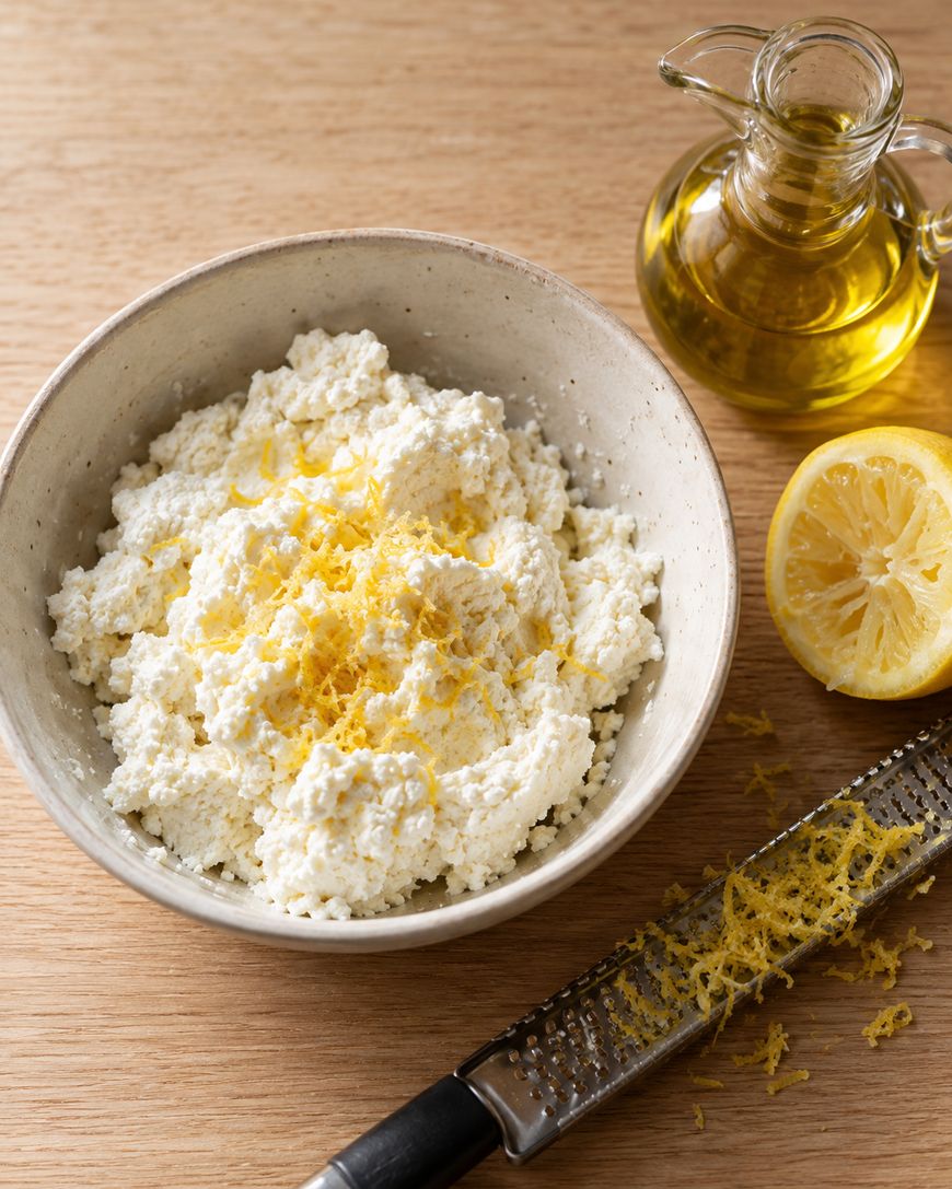 A bowl of ricotta cheese topped with lemon zest, alongside a halved lemon, a grater, and a small carafe of olive oil.