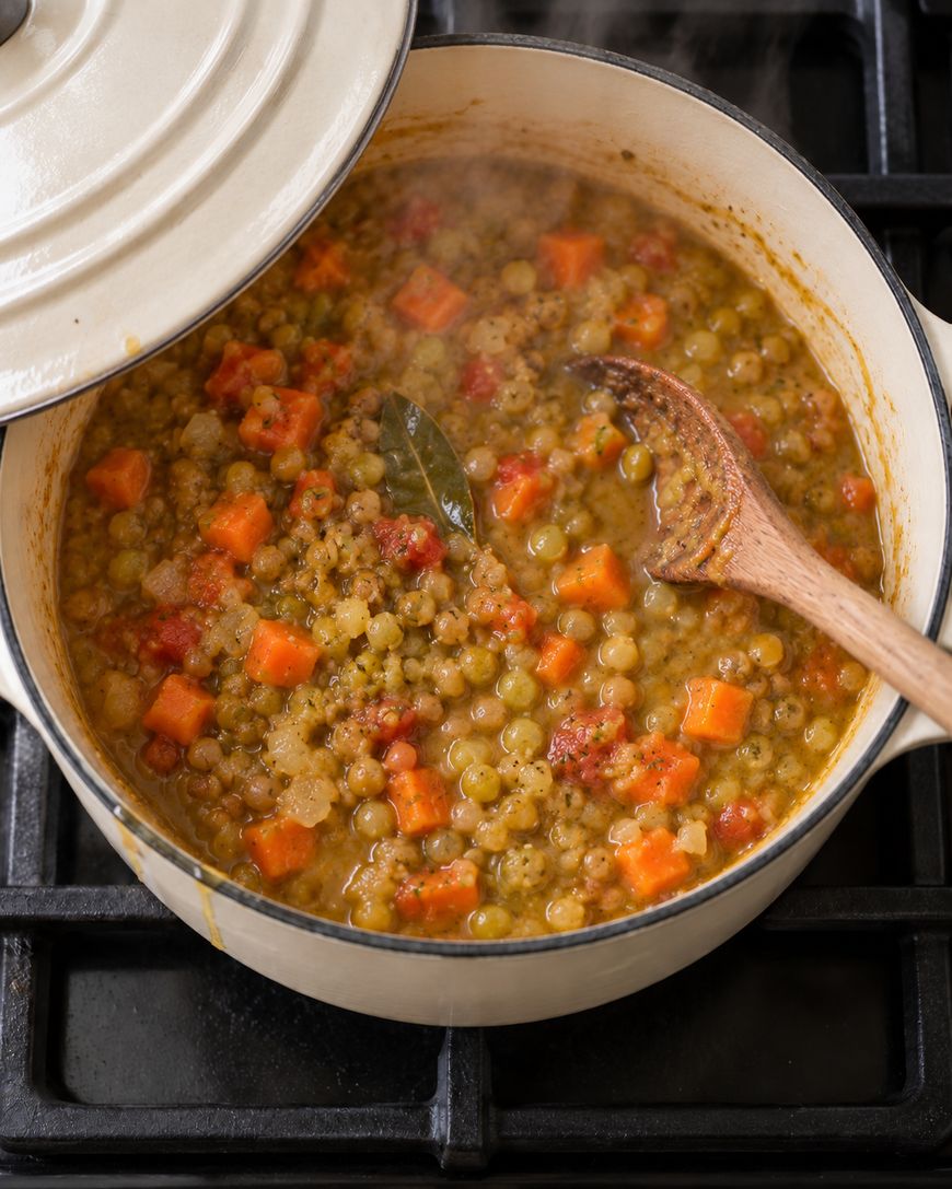 A Dutch oven filled with a thick lentil, carrot, and tomato stew with a wooden spoon and a bay leaf inside.