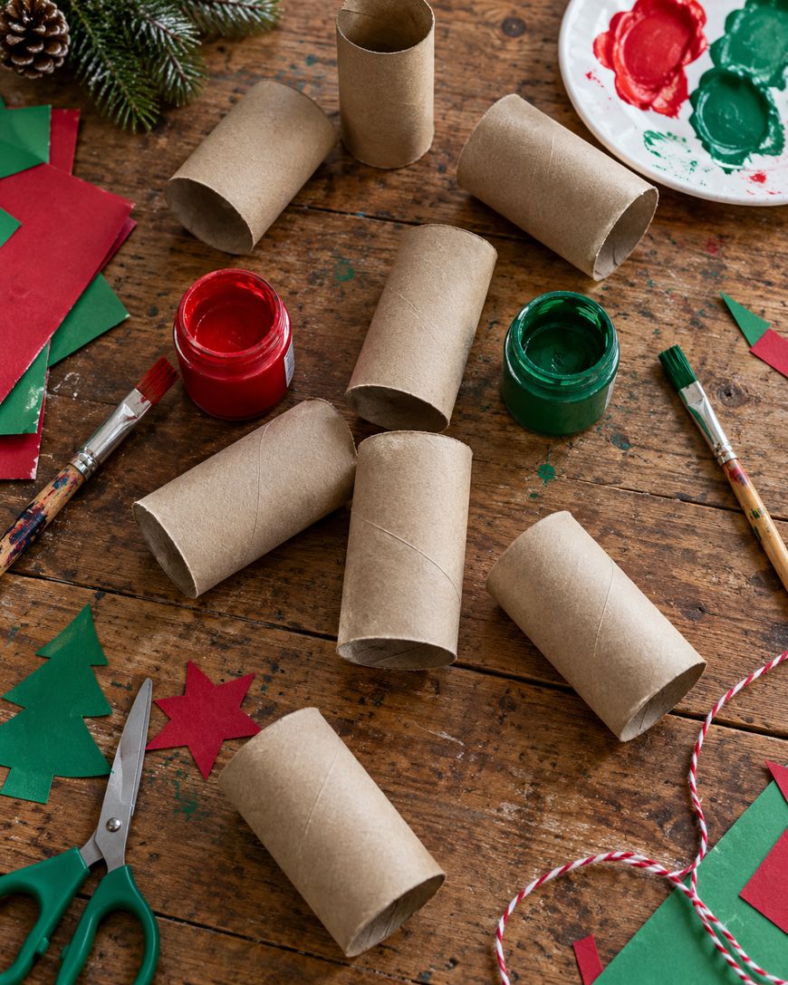 Empty cardboard tubes arranged on a wooden table with red and green paint, brushes, scissors, and paper cutouts for Christmas crafts.